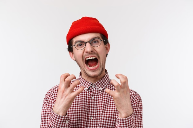 Retrato de close-up de cara hipster jovem engraçado angustiado e irritado com bigode usar gorro vermelho, óculos, apertar as mãos irritadas e incomodadas, gritando de sentimento puto