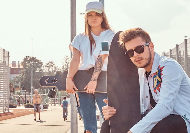 Retrato de close-up de casal jovem hipster vestido na moda com skates posando no complexo esportivo da cidade em dia ensolarado, com tons quentes.