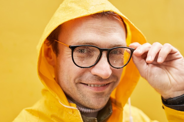 Retrato de close-up de homem caucasiano elegante vestindo capa de chuva amarela em pé contra a parede amarela tirando óculos olhando para a câmera