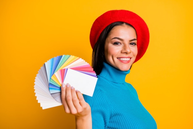 Retrato de close-up de um designer de menina morena alegre atraente e atraente segurando na mão, demonstrando cartões de paleta de diferentes tipos de cores isolados em parede de cor amarela vibrante de brilho vívido brilhante