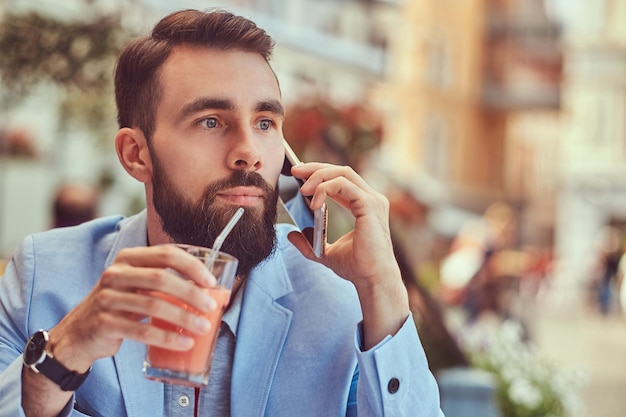 Retrato de close-up de um empresário barbudo elegante com um corte de cabelo elegante, falando por telefone, bebe um copo de suco fresco, sentado em um café ao ar livre.