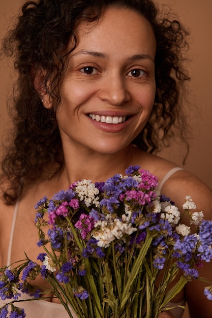 Retrato de close-up de uma encantadora mulher sorridente segurando um buquê de lindas flores silvestres, olhando para a câmera, contra um fundo bege com espaço de cópia