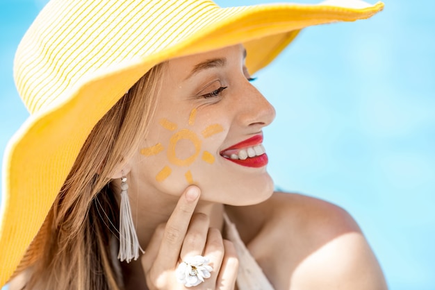 Foto retrato de close-up de uma jovem com chapéu amarelo e sol pintando sua bochecha no fundo da água azul