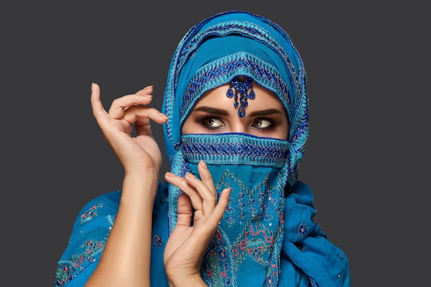 Retrato de close-up de uma jovem encantadora com belos olhos esfumaçados, vestindo um elegante hijab azul decorado com lantejoulas e joias. ela está gesticulando e desviando o olhar em um fundo escuro. hum