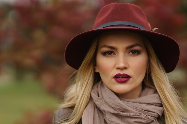 Retrato de close-up de uma linda e linda mulher com chapéu cor de vinho com pena e lenço no fundo do parque ensolarado e embaçado de outono