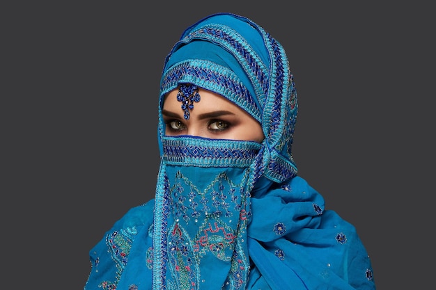 Retrato de close-up de uma linda garota com encantadores olhos esfumaçados, vestindo um hijab azul decorado com lantejoulas e joias. ela está posando de lado e olhando para a câmera em um fundo escuro. emote humano