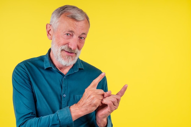 Retrato de close-up de velho feliz sênior olhando para a câmera, vestindo camisa de algodão verde com uma gola apontando o dedo para copiar spase em fundo amarelo de estúdio.