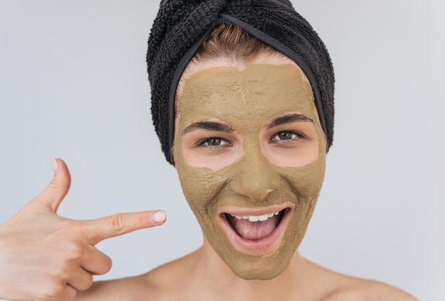 Retrato de closeup sincero de jovem feliz indica com o dedo em sua máscara orgânica de argila verde cosmética no rosto usa toalha preta no cabelo Fêmea cuida da pele do rosto isolada na parede branca