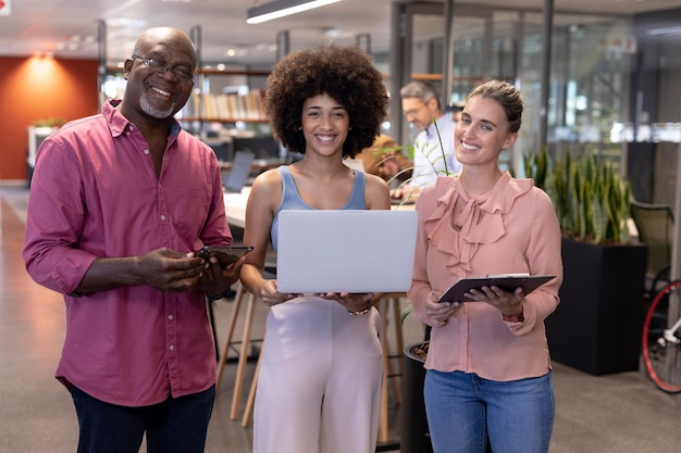 Retrato de colegas multirraciais sorridentes em pé com tecnologias sem fio no local de trabalho moderno