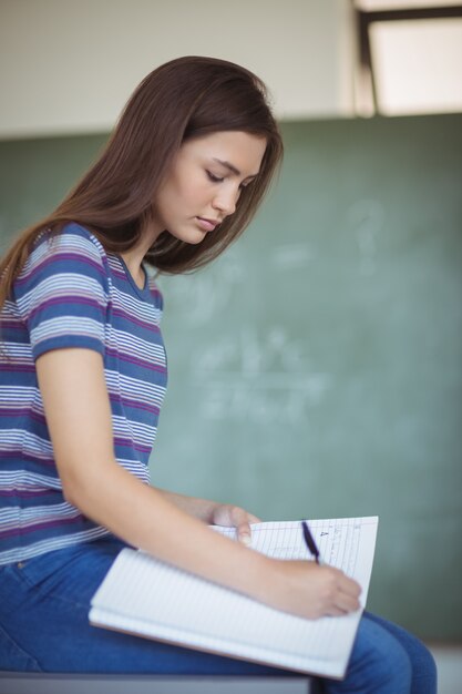 Foto retrato de colegial sentada no banco e escrevendo em um livro na sala de aula