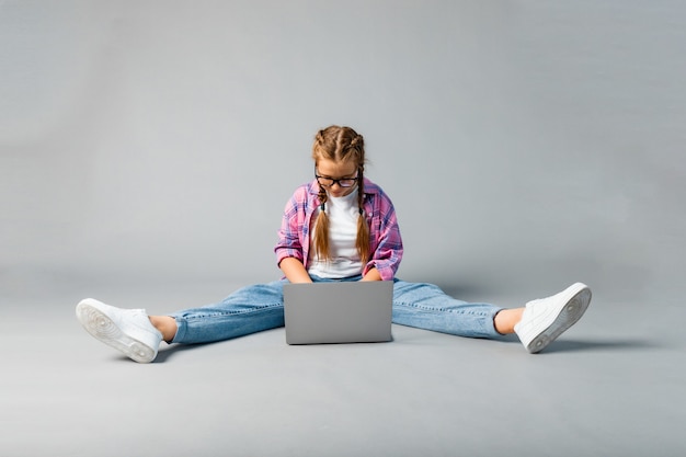 Retrato de colegial usando laptop sentado de pernas cruzadas, isolado em um fundo cinza