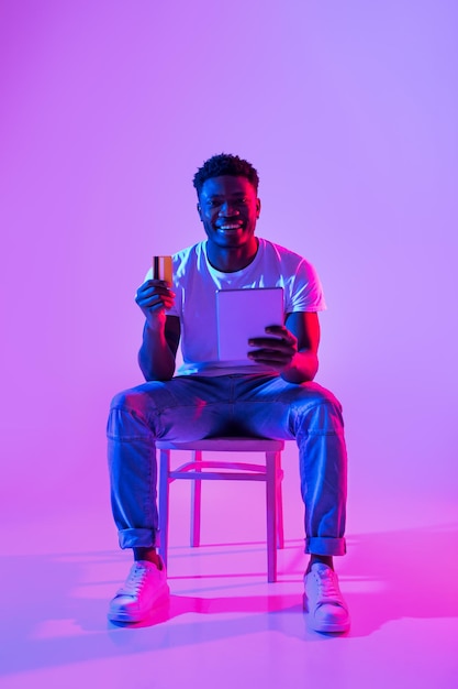 Retrato de compras online de um negro alegre com laptop e cartão de crédito sentado na cadeira em neon