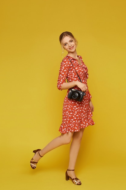 Retrato de comprimento total de uma garota modelo em um vestido de verão colorido, posando com uma câmera vintage sobre yel.