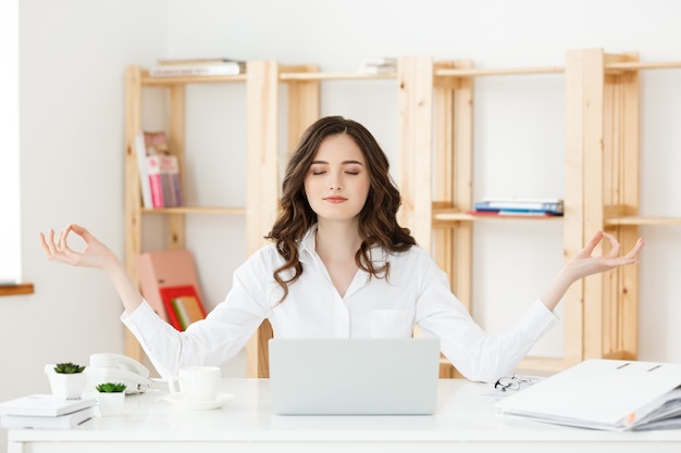 Retrato de conceito de saúde e negócios jovem perto do laptop praticando meditação na mesa do escritório em frente a aulas de ioga online com laptop fazendo uma pausa por um minuto