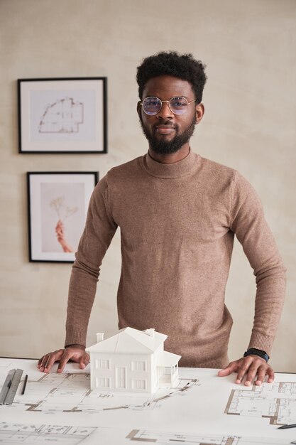 Retrato de conteúdo jovem arquiteto de construção afro-americano em copos em pé à mesa com planta baixa e modelo D de casa