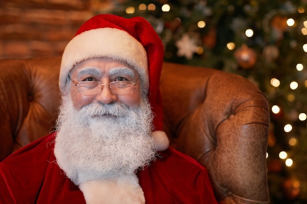 Retrato de conteúdo sênior de Papai Noel no boné vermelho e óculos, sentado na poltrona contra o Natal ...