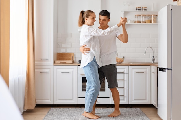 Retrato de corpo inteiro de casal feliz comemorando a compra de uma casa nova, dançando contra o conjunto de cozinha branca, expressando emoções românticas positivas, vestindo roupas de estilo casual.