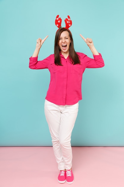 Retrato de corpo inteiro de diversão jovem em blusa de camisa rosa, calça branca, chifres de veado posando isolados na parede pastel azul rosa brilhante.