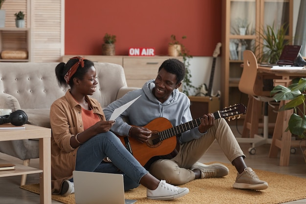 Retrato de corpo inteiro de dois jovens músicos afro-americanos tocando violão e escrevendo música juntos, sentados no chão em um estúdio de gravação, copie o espaço