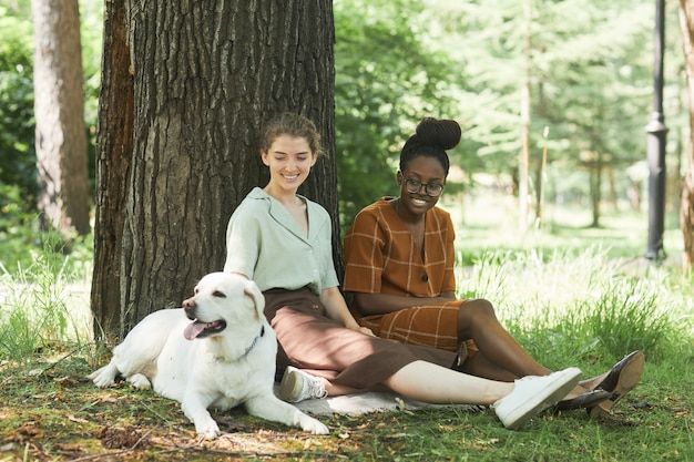 Retrato de corpo inteiro de duas mulheres sentadas na grama verde no parque com um cachorro Labrador sorrindo, copie o espaço