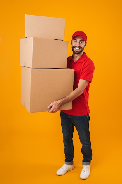 Retrato de corpo inteiro de entregador alegre em uniforme vermelho, sorrindo enquanto carregava caixas de embalagem isoladas sobre amarelo
