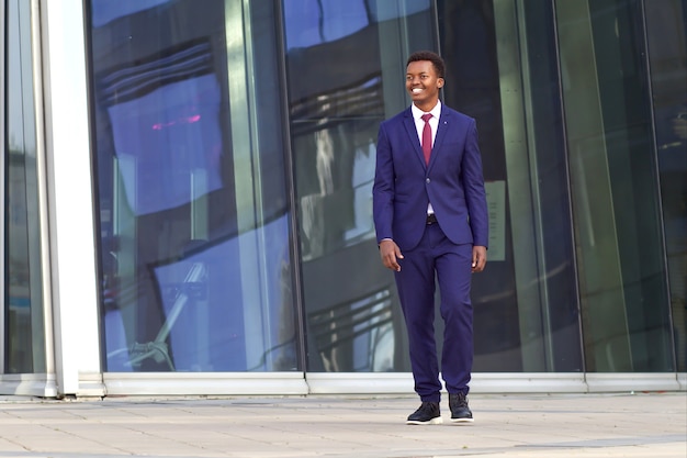 Retrato de corpo inteiro de feliz positivo negro afro-americano jovem empresário em terno formal indo e sorrindo. Escritório comercial masculino ao ar livre