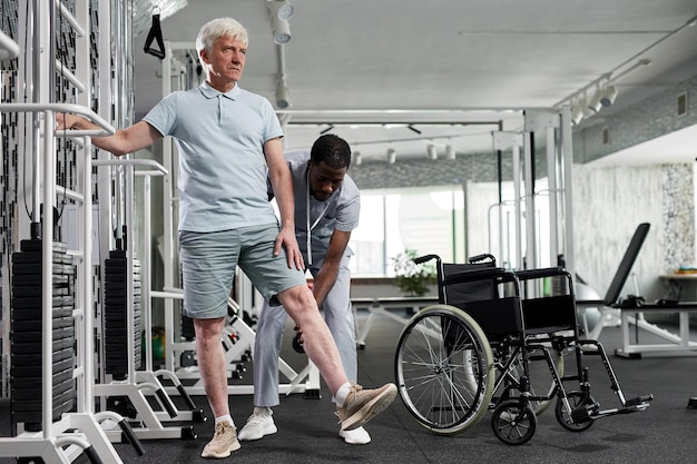 Retrato de corpo inteiro de homem sênior fazendo exercícios de reabilitação no ginásio com espaço de cópia assistente