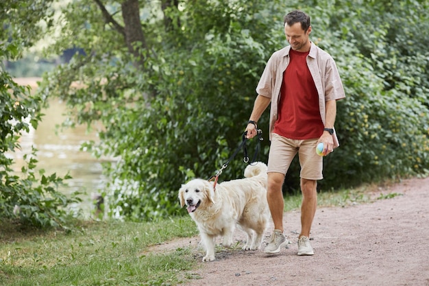 Retrato de corpo inteiro de homem sorridente andando com cachorro ao ar livre no verão no parque verde