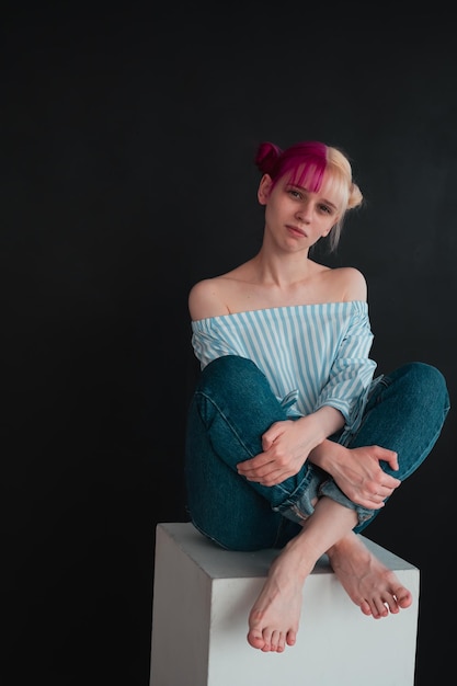 Retrato de corpo inteiro de jovem com cabelo branco e rosa, sentado no cubo branco sobre fundo preto