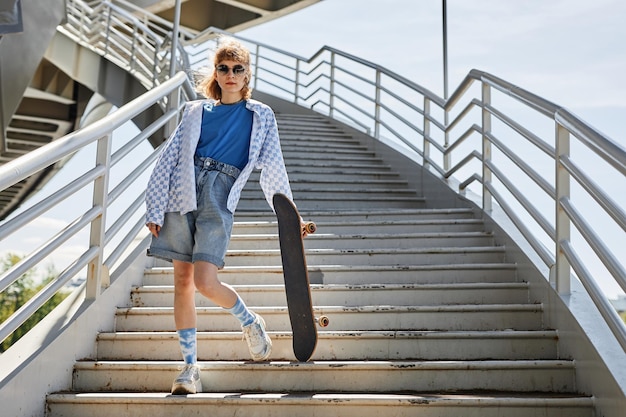 Retrato de corpo inteiro de jovem com skate posando ao ar livre na luz do sol e em pé na escada