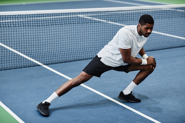 Retrato de corpo inteiro de jovem desportista negro esticando e aquecendo na quadra de tênis coberta