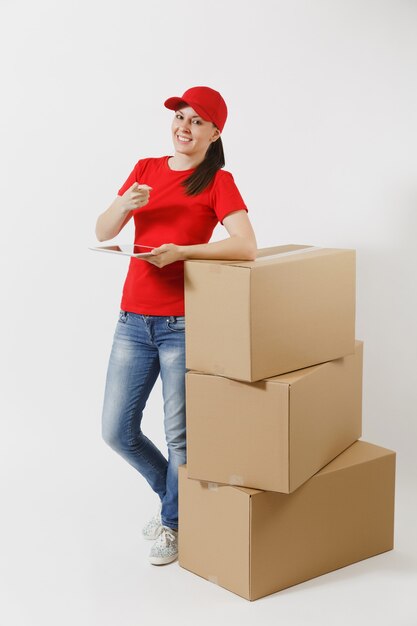 Retrato de corpo inteiro de jovem entrega em boné vermelho, t-shirt isolado no fundo branco. Correio feminino em pé perto de caixas de papelão vazias com computador tablet pc. Recebendo pacote. Copie o espaço