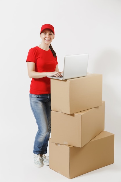 Retrato de corpo inteiro de jovem entrega em boné vermelho, t-shirt isolado no fundo branco. Correio feminino em pé perto de caixas de papelão vazias com o computador laptop pc. Recebendo pacote. Copie o espaço