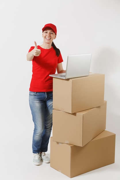 Retrato de corpo inteiro de jovem entrega em boné vermelho, t-shirt isolado no fundo branco. Correio feminino em pé perto de caixas de papelão vazias com o computador laptop pc. Recebendo pacote. Copie o espaço