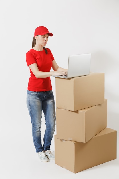 Retrato de corpo inteiro de jovem entrega em boné vermelho, t-shirt isolado no fundo branco. Correio feminino em pé perto de caixas de papelão vazias com o computador laptop pc. Recebendo pacote. Copie o espaço