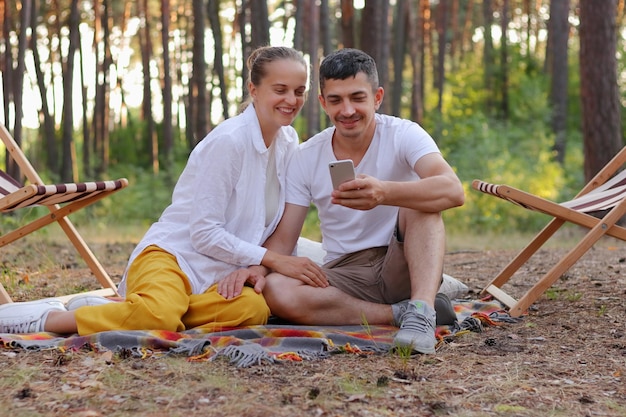 Retrato de corpo inteiro de lindo casal jovem sentado em uma manta na floresta e usando o celular navegando na internet ou compartilhando fotos de suas férias na natureza