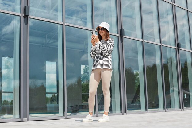 Retrato de corpo inteiro de mulher caucasiana vestindo camisa listrada panamá e óculos de sol usando smartphone posando perto de prédio com grandes janelas fazendo selfie