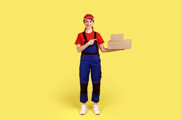 Retrato de corpo inteiro de mulher de correio amigável sorridente apontando para pacotes de papelão em suas mãos serviço de entrega rápida usa macacão e boné vermelho Estúdio interior isolado em fundo amarelo