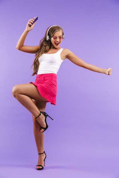 Retrato de corpo inteiro de mulher enérgica feliz em roupas de verão e óculos de sol dançando enquanto ouve música através de fones de ouvido sem fio,