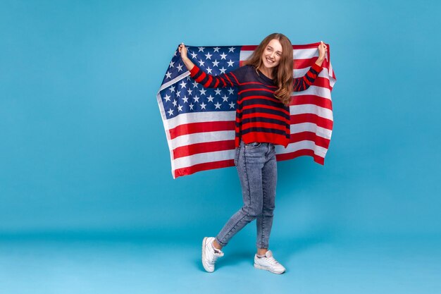 Retrato de corpo inteiro de mulher feliz no suéter listrado estilo casual, segurando a bandeira dos EUA e olhando para a câmera com sorriso comemorando feriados nacionais Tiro de estúdio interior isolado em fundo azul