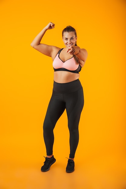 Foto retrato de corpo inteiro de mulher gordinha esportiva em sutiã esportivo mostrando seu bíceps, isolado sobre fundo amarelo