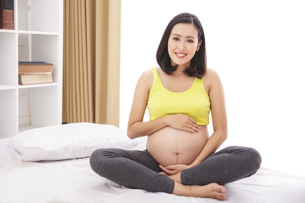 Retrato de corpo inteiro de mulher grávida asiática sorrindo enquanto está sentada na cama