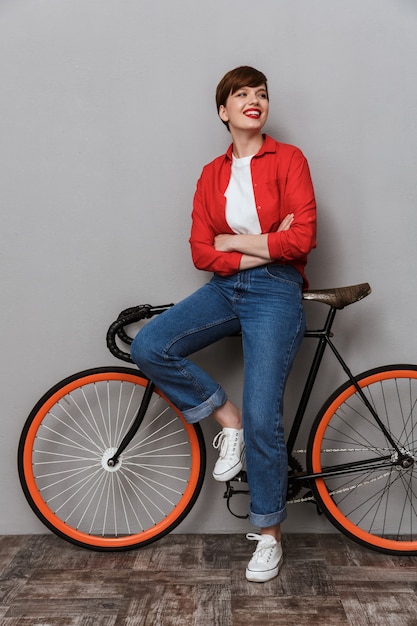 Retrato de corpo inteiro de mulher satisfeita, sorrindo e em pé ao lado de uma bicicleta elegante isolada sobre uma parede cinza