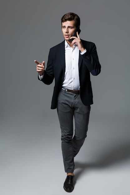 Retrato de corpo inteiro de um belo jovem empresário vestido com um terno isolado sobre uma parede cinza, caminhando, falando no celular, apontando