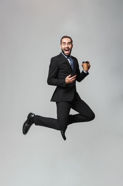 Foto retrato de corpo inteiro de um empresário bonito e confiante vestindo terno isolado, segurando café para viagem, usando telefone celular, pulando