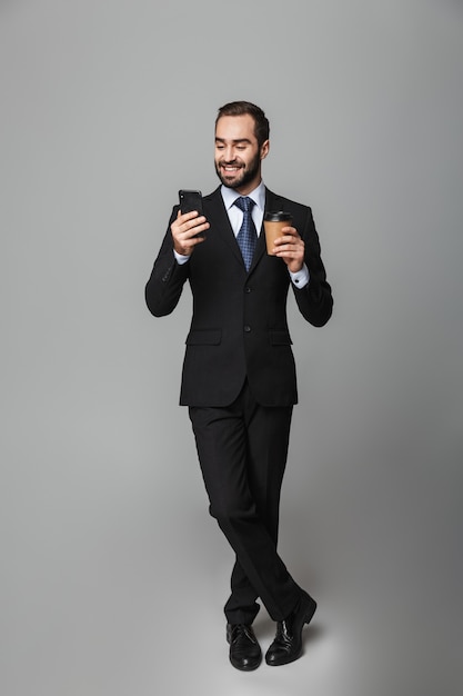 Retrato de corpo inteiro de um empresário bonito e confiante vestindo terno isolado, segurando café para viagem, usando telefone celular