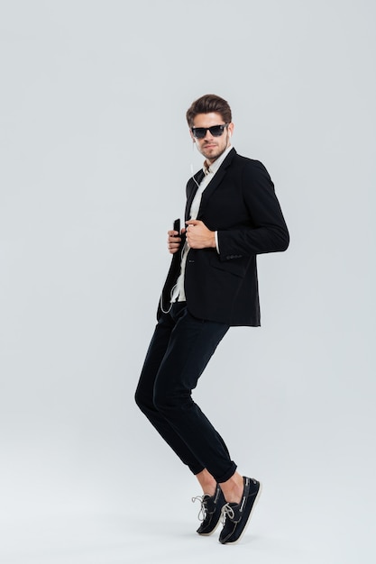 Retrato de corpo inteiro de um empresário sério ouvindo música com fone de ouvido e na ponta dos pés sobre uma parede cinza