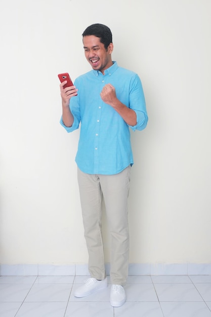 Retrato de corpo inteiro de um homem adulto asiático com os punhos cerrados, mostrando uma expressão animada enquanto olha para seu telefone celular