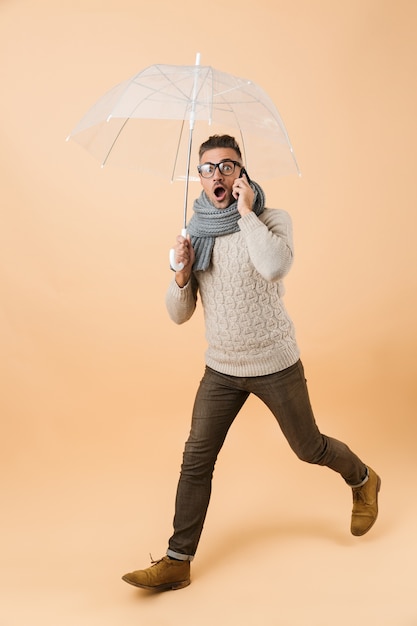 Retrato de corpo inteiro de um homem chocado com um suéter e um lenço andando sob o guarda-chuva isolado sobre uma parede bege, falando no celular