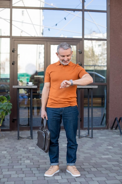 Retrato de corpo inteiro de um homem com uma bolsa de couro do lado de fora do café e olhando para o relógio de pulso